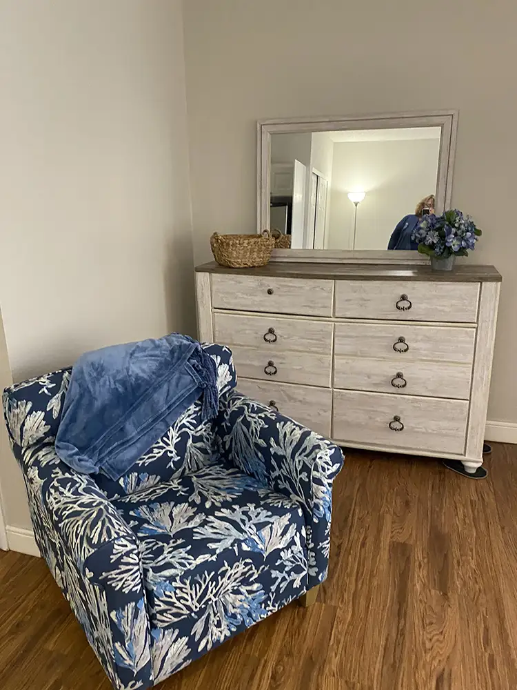 Cozy resident room corner with a blue patterned armchair and white dresser at Springwood Court in Florida.